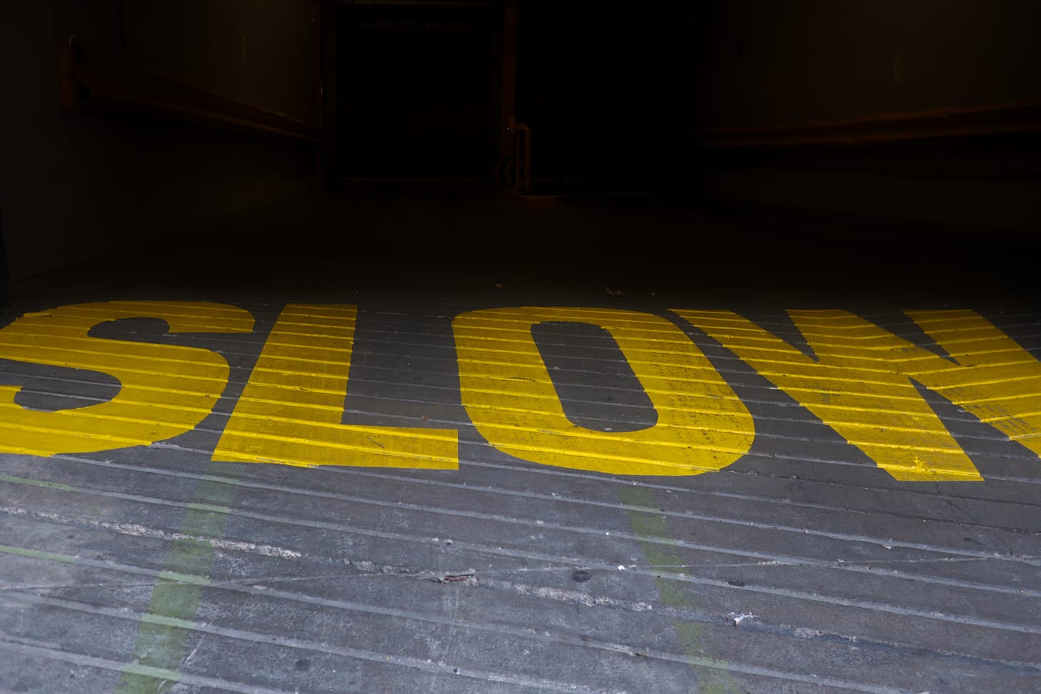 The word “SLOW” painted in vivid yellow on a driveway. The road ahead is filled with deep shadows.